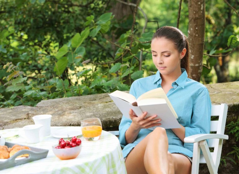 A beautiful girl, enjoying a healthy breakfast outside on a sunny day, reads a book, understanding the benefits of daily routines.