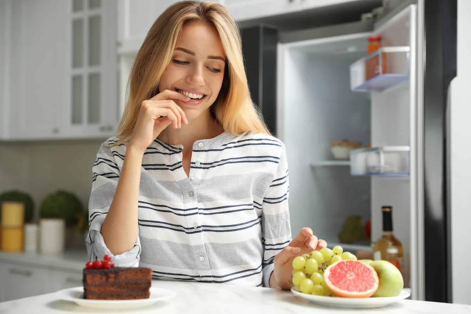 In her decluttered and organized kitchen, a woman deliberates between choosing cake and healthy fruits on the table, leaning towards the healthier option.