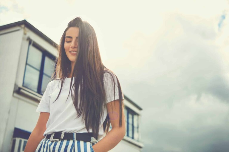 Highly sensitive woman wearing striped pants puts hand in pocket and smiles as hair hangs in face giving the impression that she is shy.