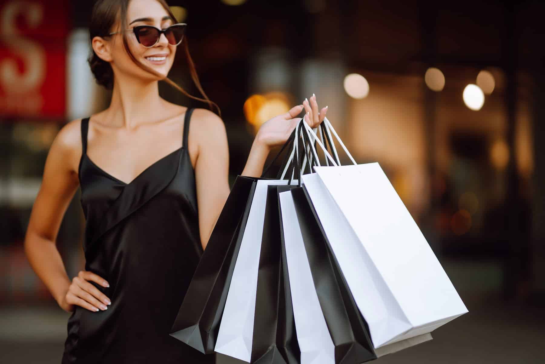 A woman dressed in a black dress is holding black and white shopping bags, visibly enjoying her shopping spree, but perhaps overindulging by purchasing items she doesn't actually need.