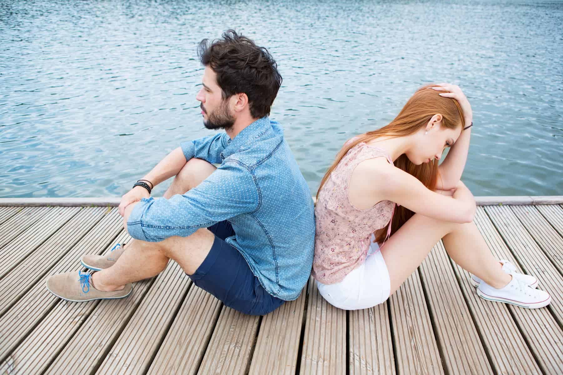 Highly sensitive couple seated by the water with their backs against each other, contemplating the challenges of relationships.