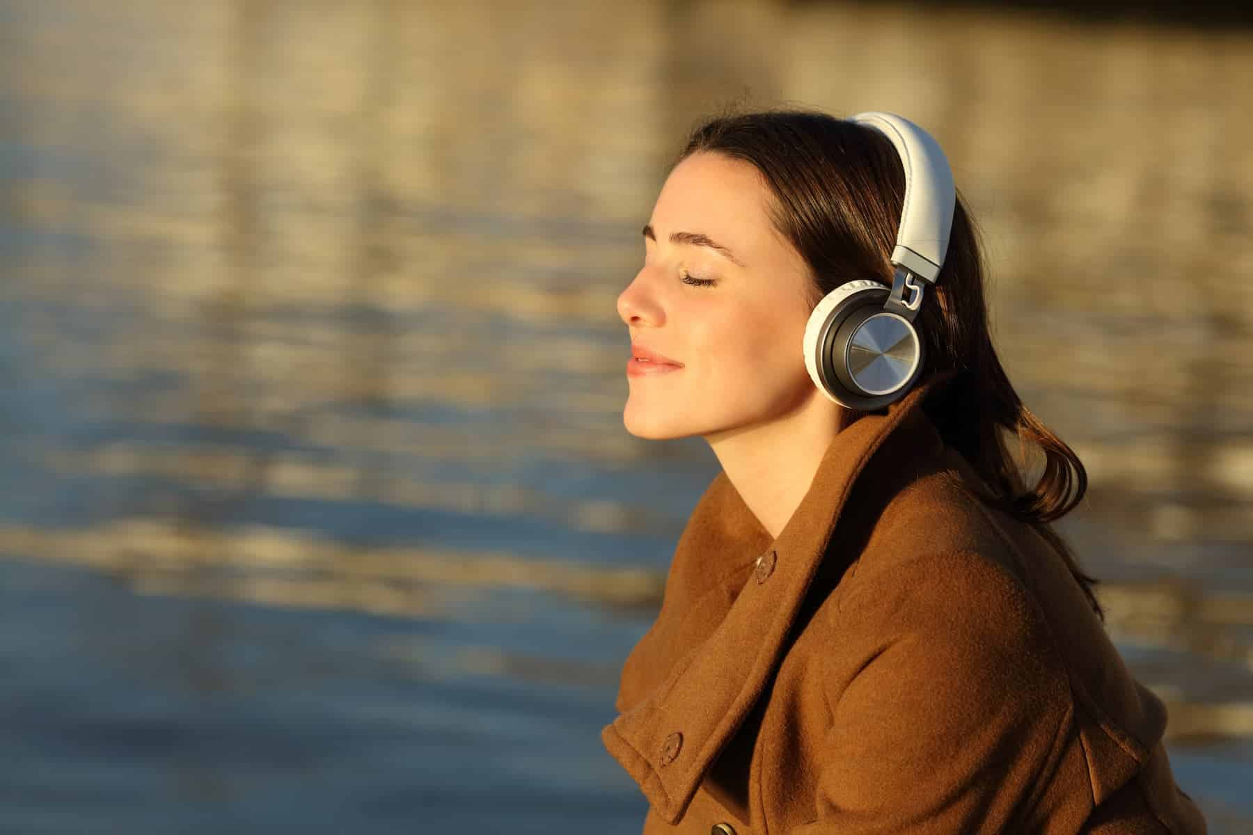 A woman, dressed in a brown coat, stands by the lake with her eyes closed, wearing headphones, as she visualizes her goals and smiles to herself.