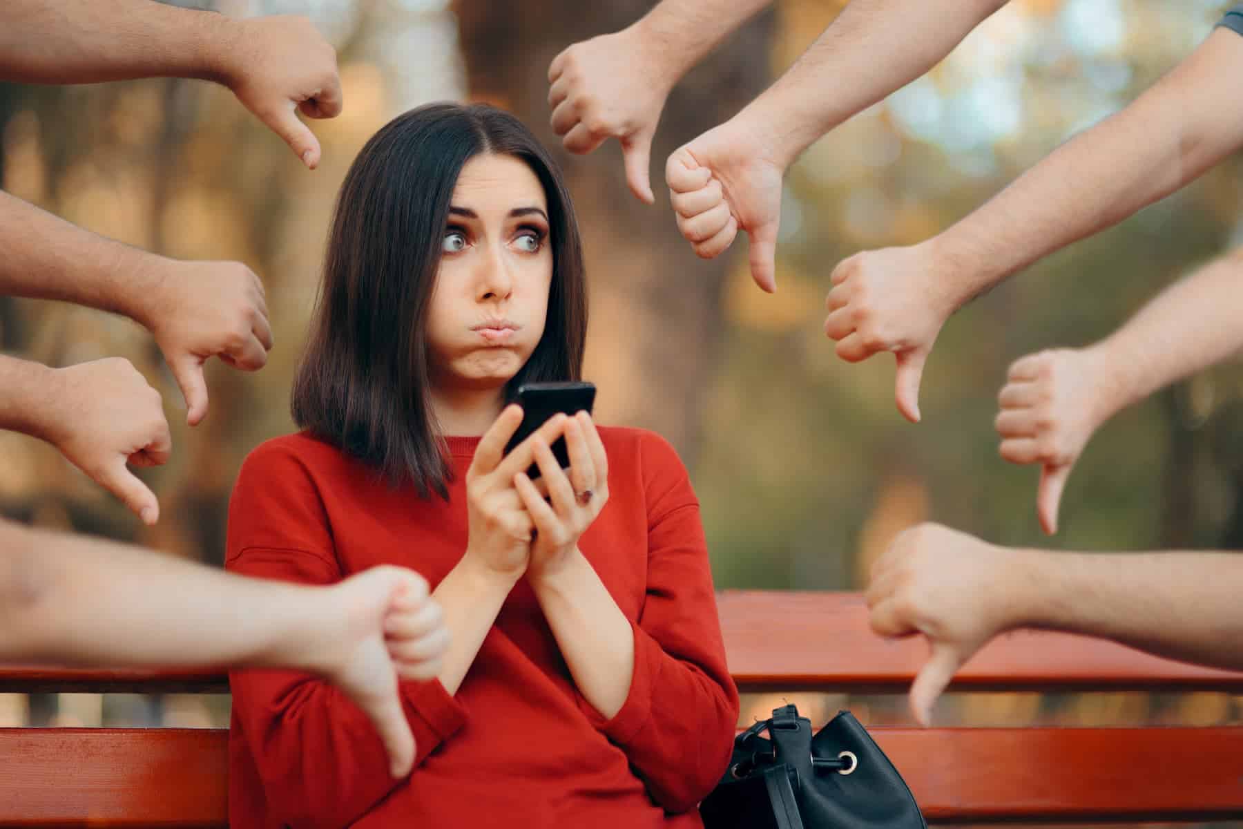 Awoman in a red sweater sitting on a park bench, looking a bit let down. She's an online influencer, checking out the feedback she's getting.