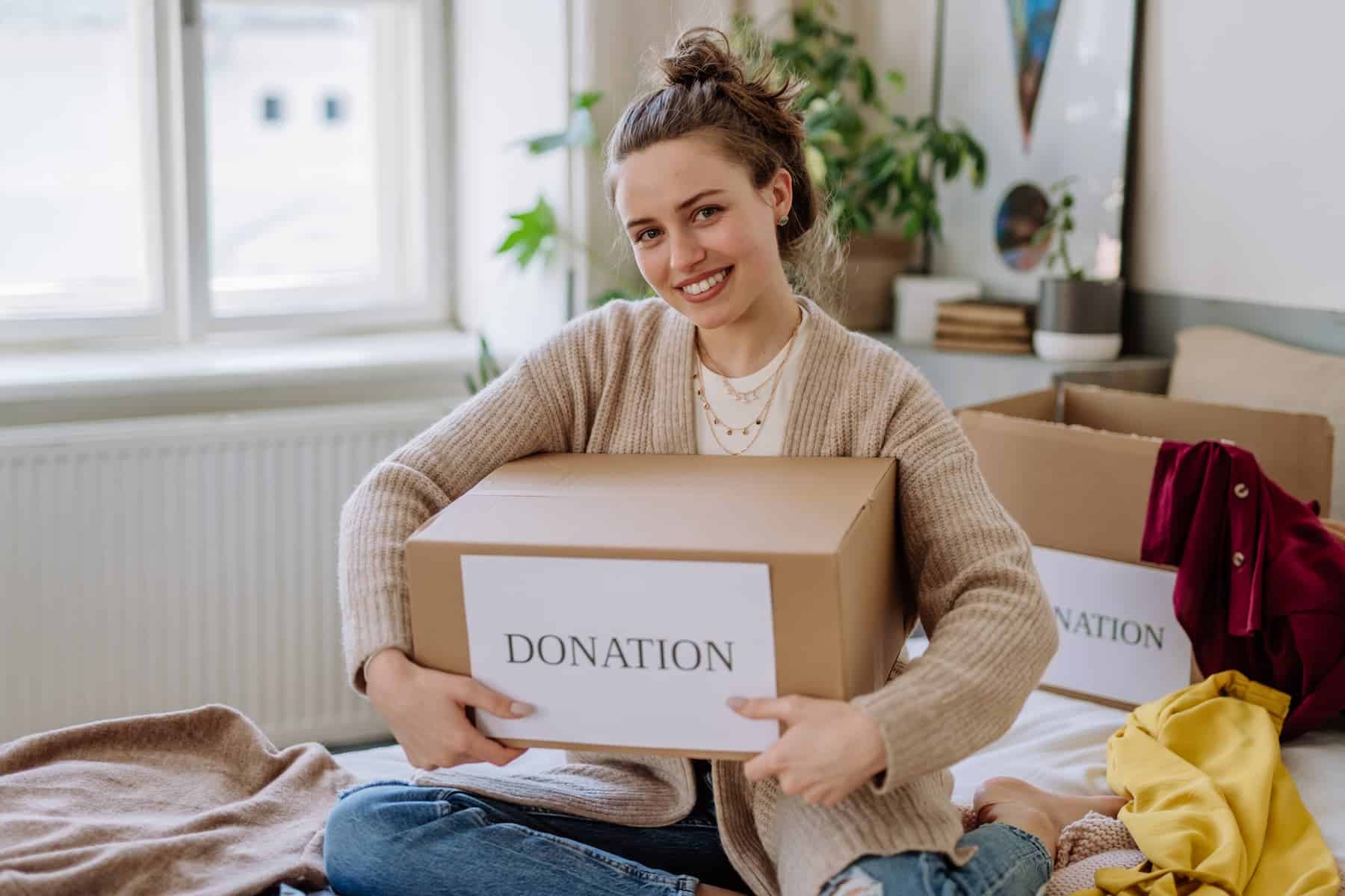 A young woman sits on her bed, carefully packing clothes into a donation box — embracing the idea: clear your space, clear your mind.