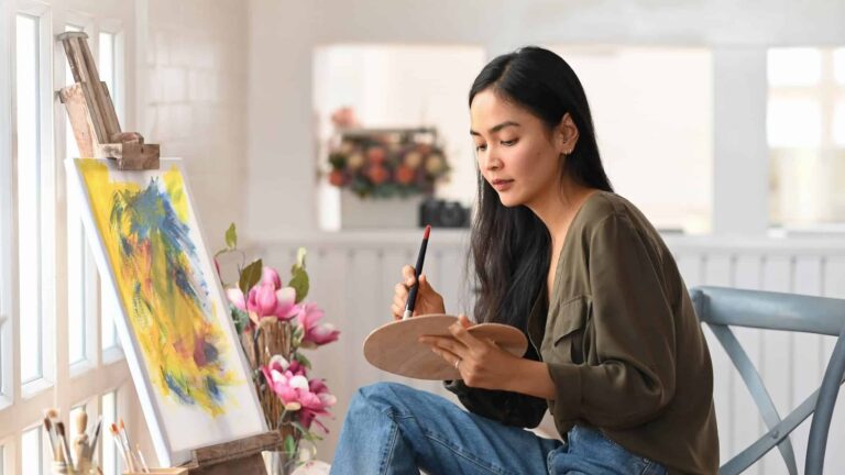 A young woman sits in her studio, painting with focused determination, enjoying new creative hobbies to express herself.