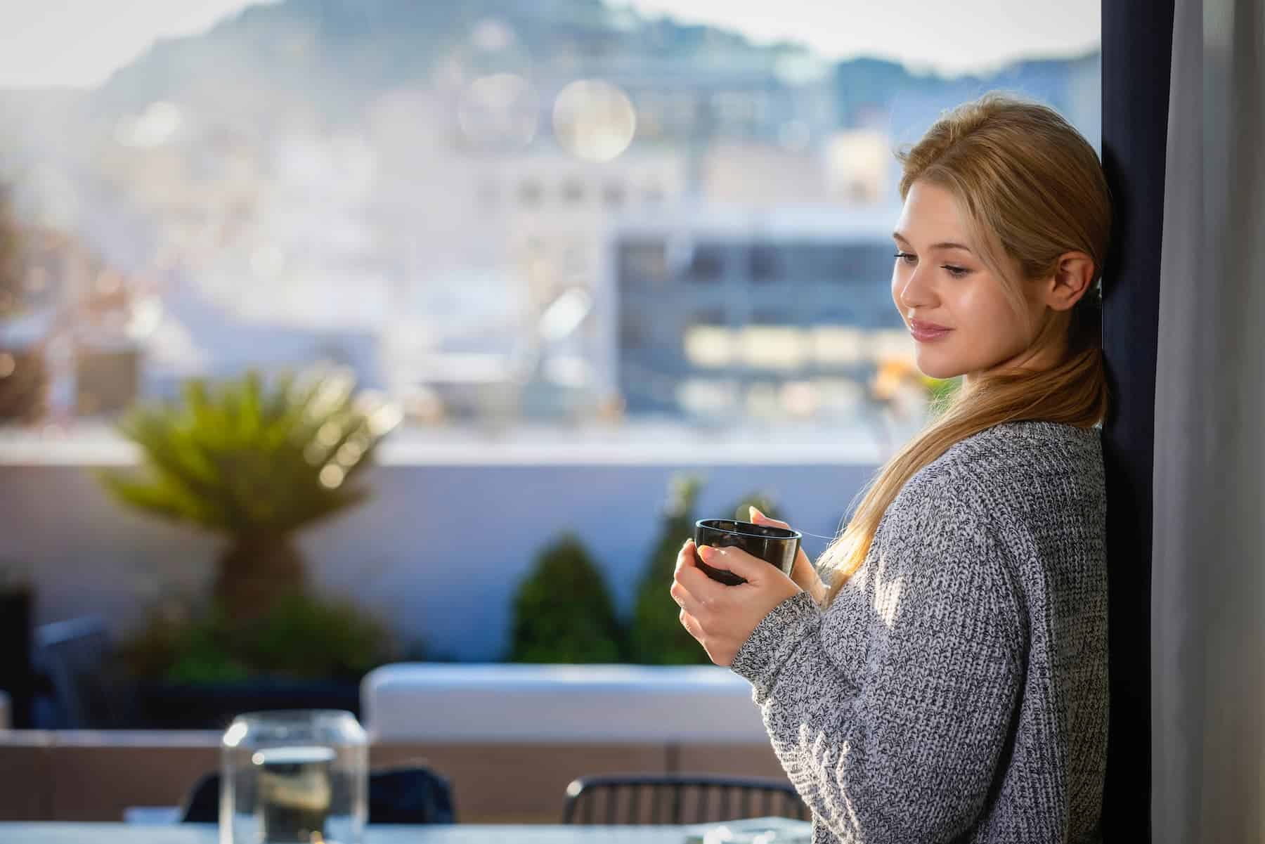 A woman sips coffee at home, gazing out her large window, enjoying every moment after getting some great daily routine tips from a friend.