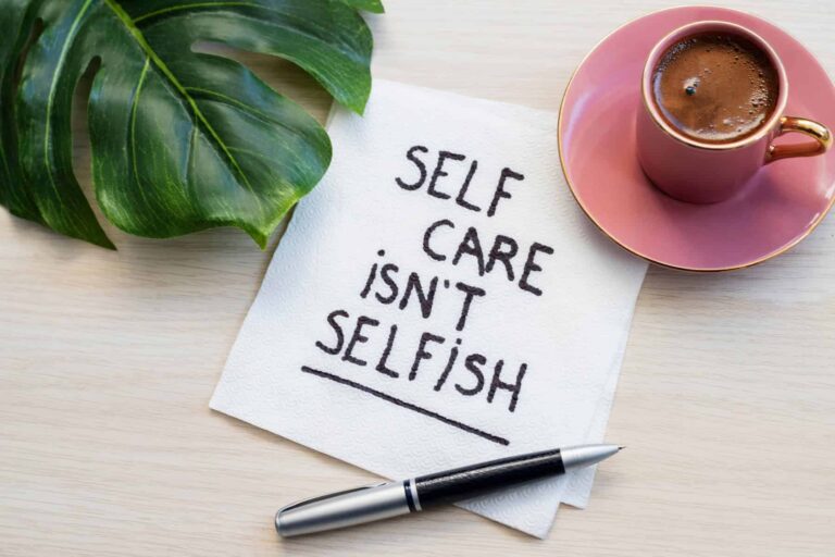 An inspirational reminder on a napkin next to a pen and a pink coffee cup: 'Self-care practices are not selfish.'