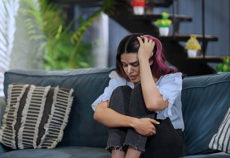 An upset young woman sitting on a sofa is noticing symptoms of the dark night of the soul.