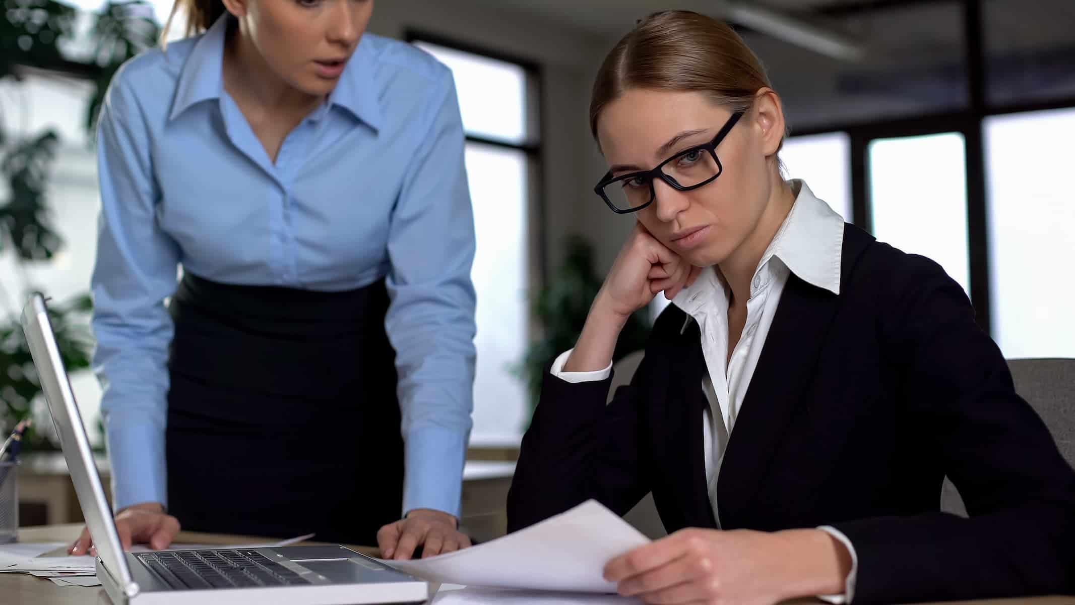 A woman leans on her hand at her desk while reviewing papers as another person stands close, illustrating pressure linked to dealing with criticism as a highly sensitive person.