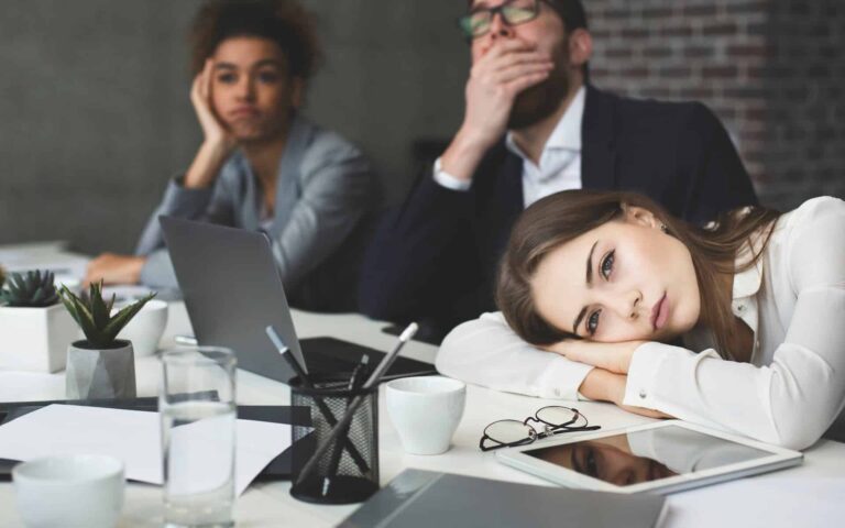 Three business people appearing bored, with one woman resting her head on the table not understanding why they can never escape mediocrity.