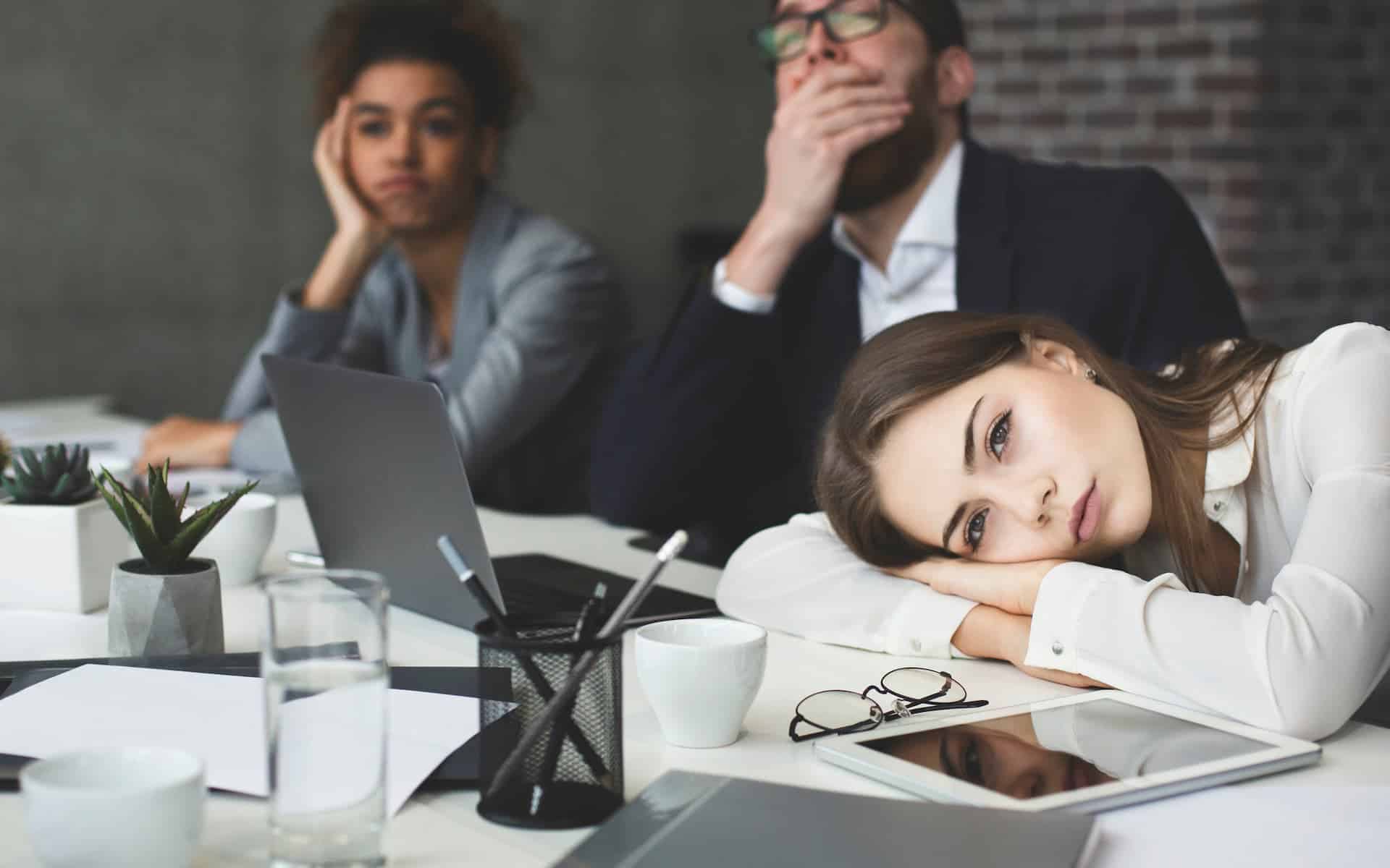 Three business people appearing bored, with one woman resting her head on the table not understanding why they can never escape mediocrity.