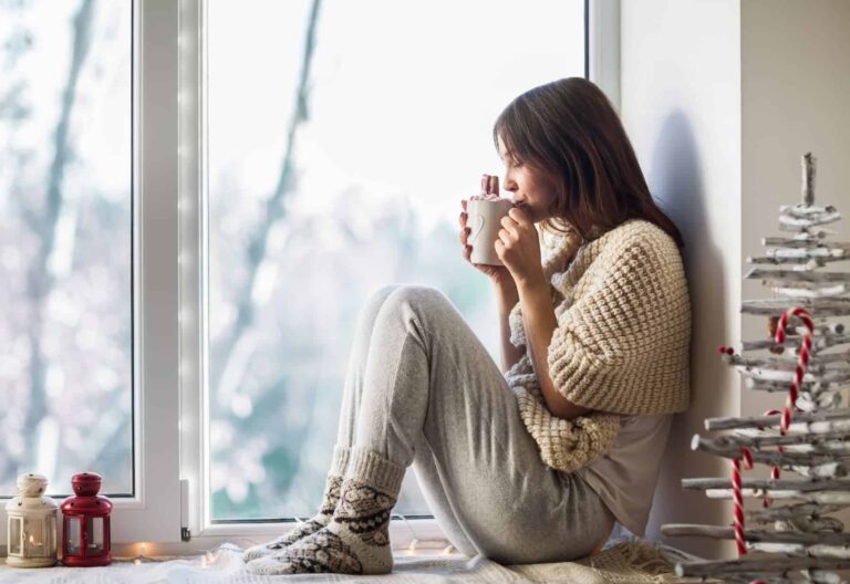 Woman by a window with a mug, reflecting on getting out of the comfort zone.