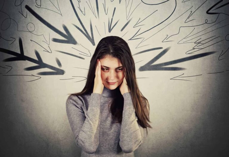 A highly sensitive woman in an insensitive world stands with her hands on her temples against a grey wall, with black painted arrows pointing at her face.