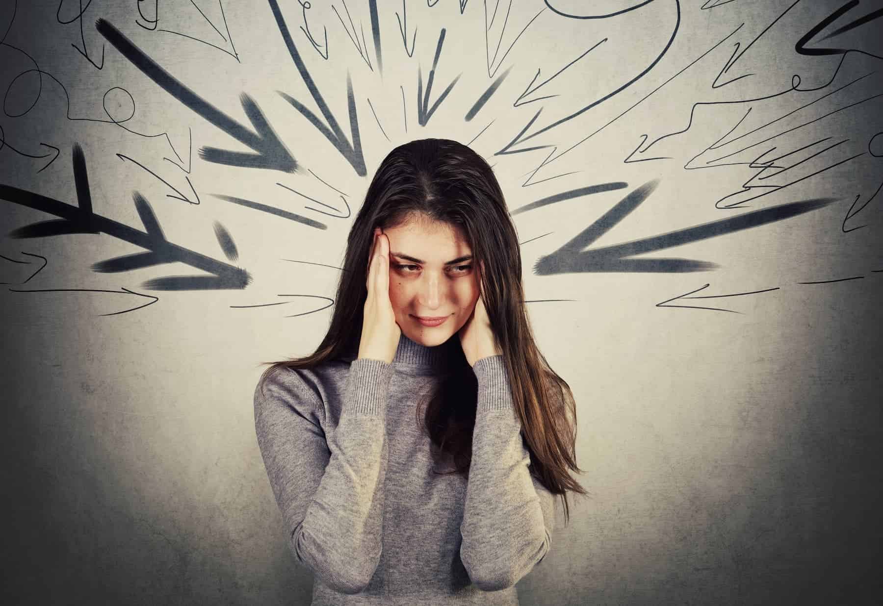 A highly sensitive woman in an insensitive world stands with her hands on her temples against a grey wall, with black painted arrows pointing at her face.