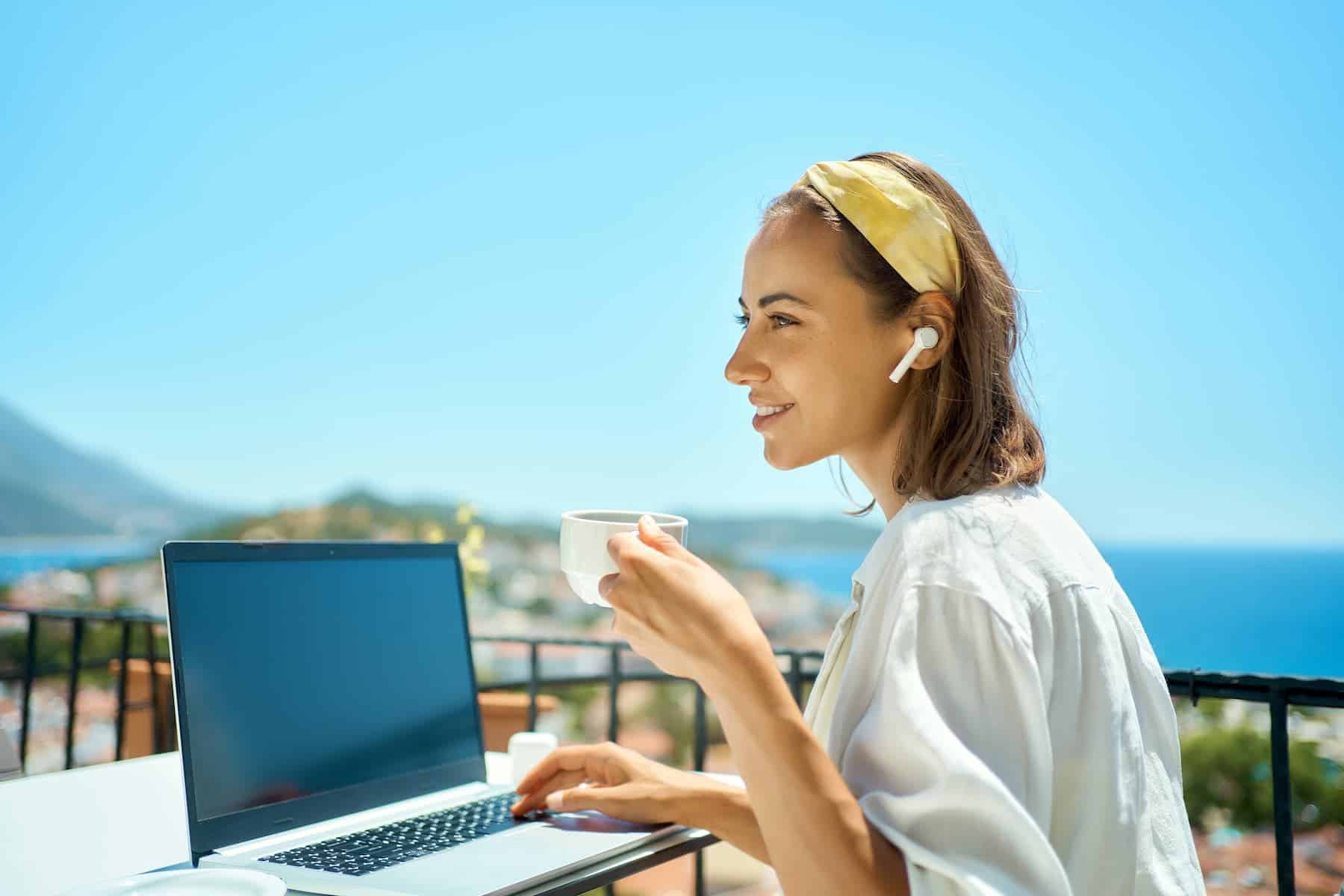 Woman working on her laptop in her balcony and enjoying a cup of coffee in the beautiful weather knows how to balance work and life.