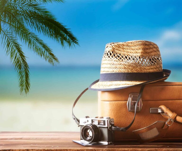 A vintage suitcase, hipster hat, photo camera, and passport on a wooden deck with the beach in the background on a sunny day, showcasing how to reduce stress while traveling.