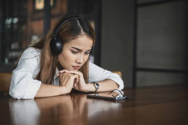 A woman wearing headphones rests her chin on her hands while studying her phone.