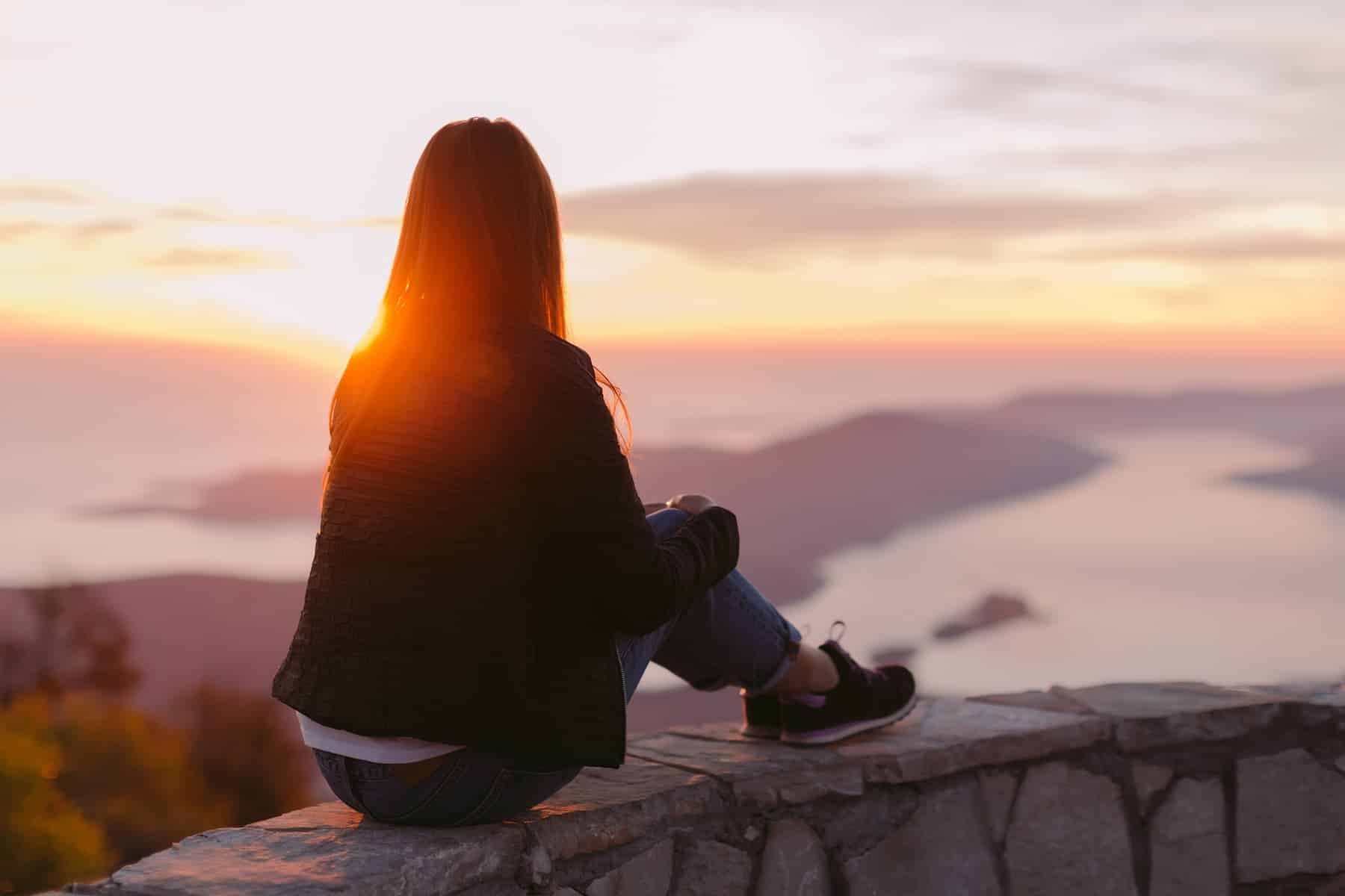 A woman sits on a stone wall overlooking a body of water at sunset, enjoying a quiet, introspective moment that reflects her introverted nature.