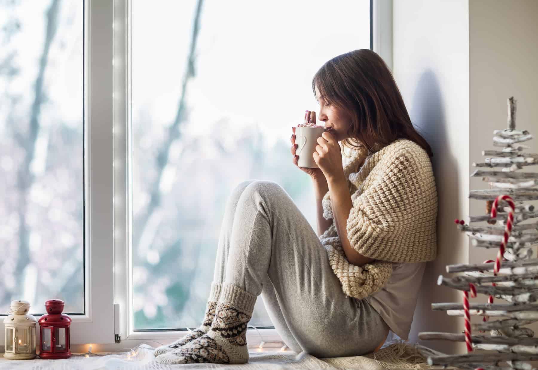 A woman hesitates to confront her mediocre mindset, finding solace in her comfort zone as she sits on the window sill, drinking hot coffee.