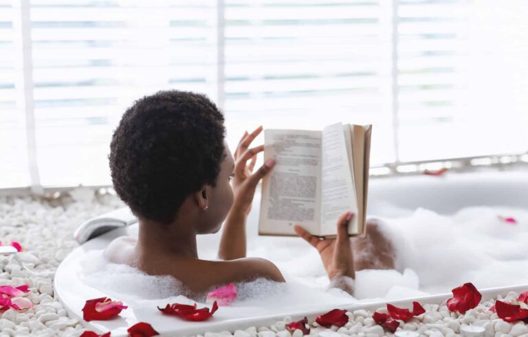 A young woman relaxes in a hot tub filled with rose petals, reading a book — practicing negative self-care by completely ignoring her other responsibilities.