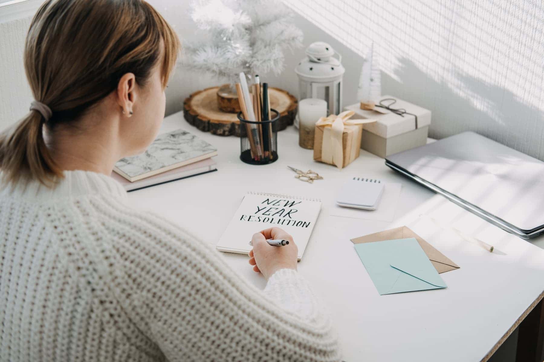A woman in a white sweater sits at a table, writing New Year's resolution in an open notepad.