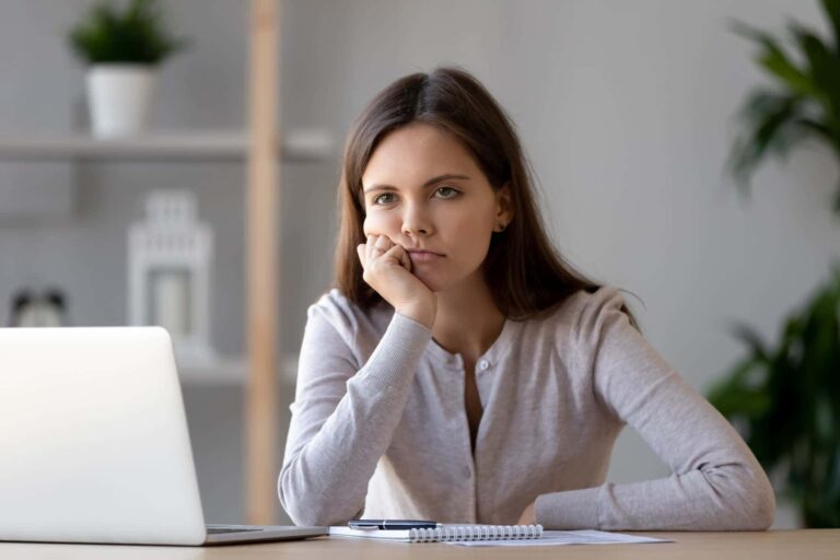 A woman sits at a desk with a laptop and notebook, resting her chin on her hand and looking bored.