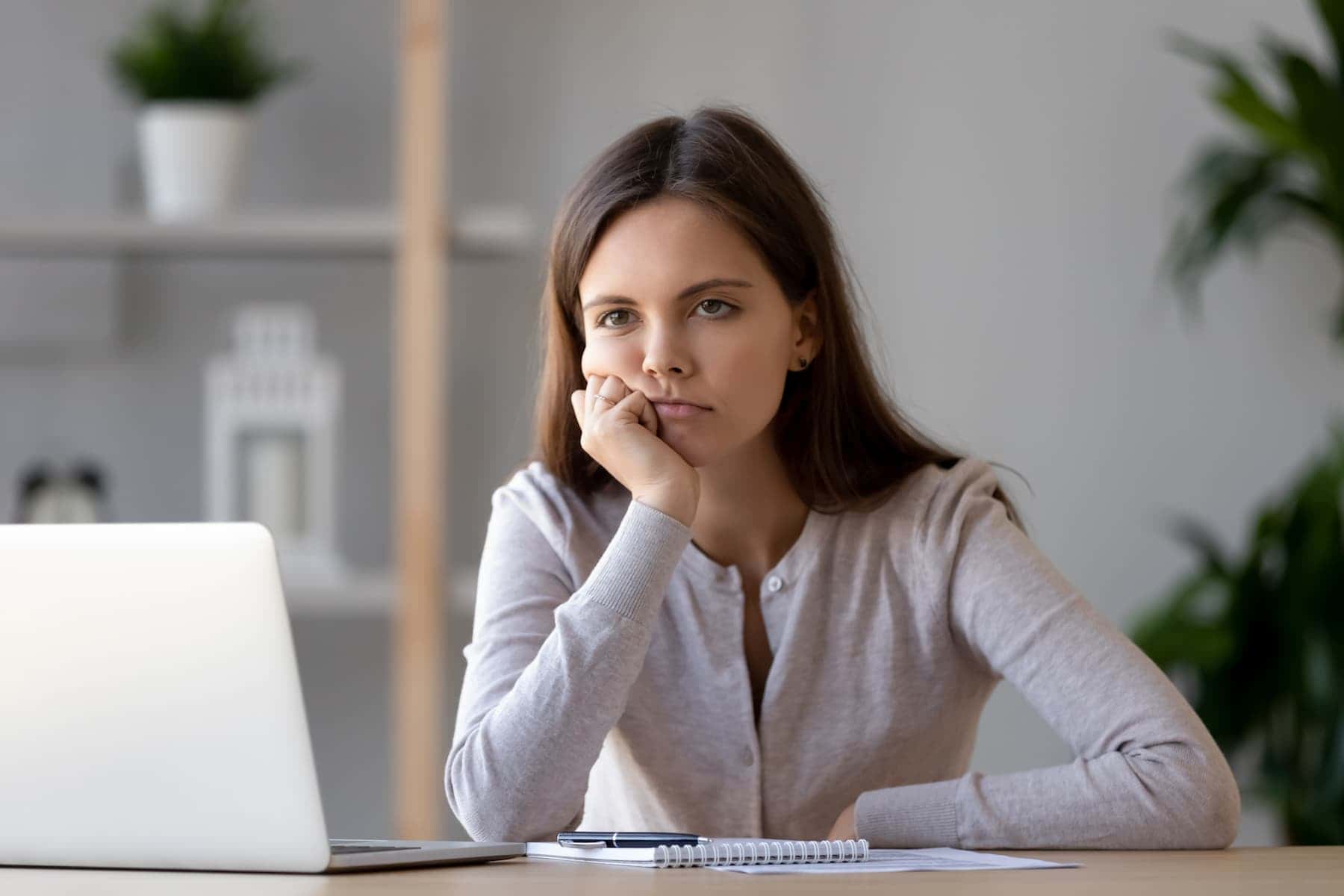 A woman sits at a desk with a laptop and notebook, resting her chin on her hand and looking bored.