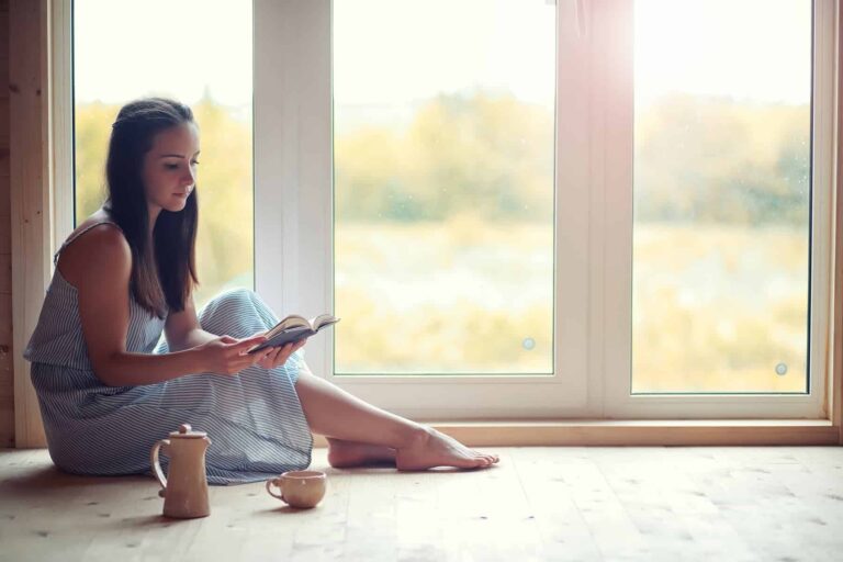 A young woman finds solace by the window, immersed in a book and sipping tea — she knows plenty of self-care ideas for unwinding.