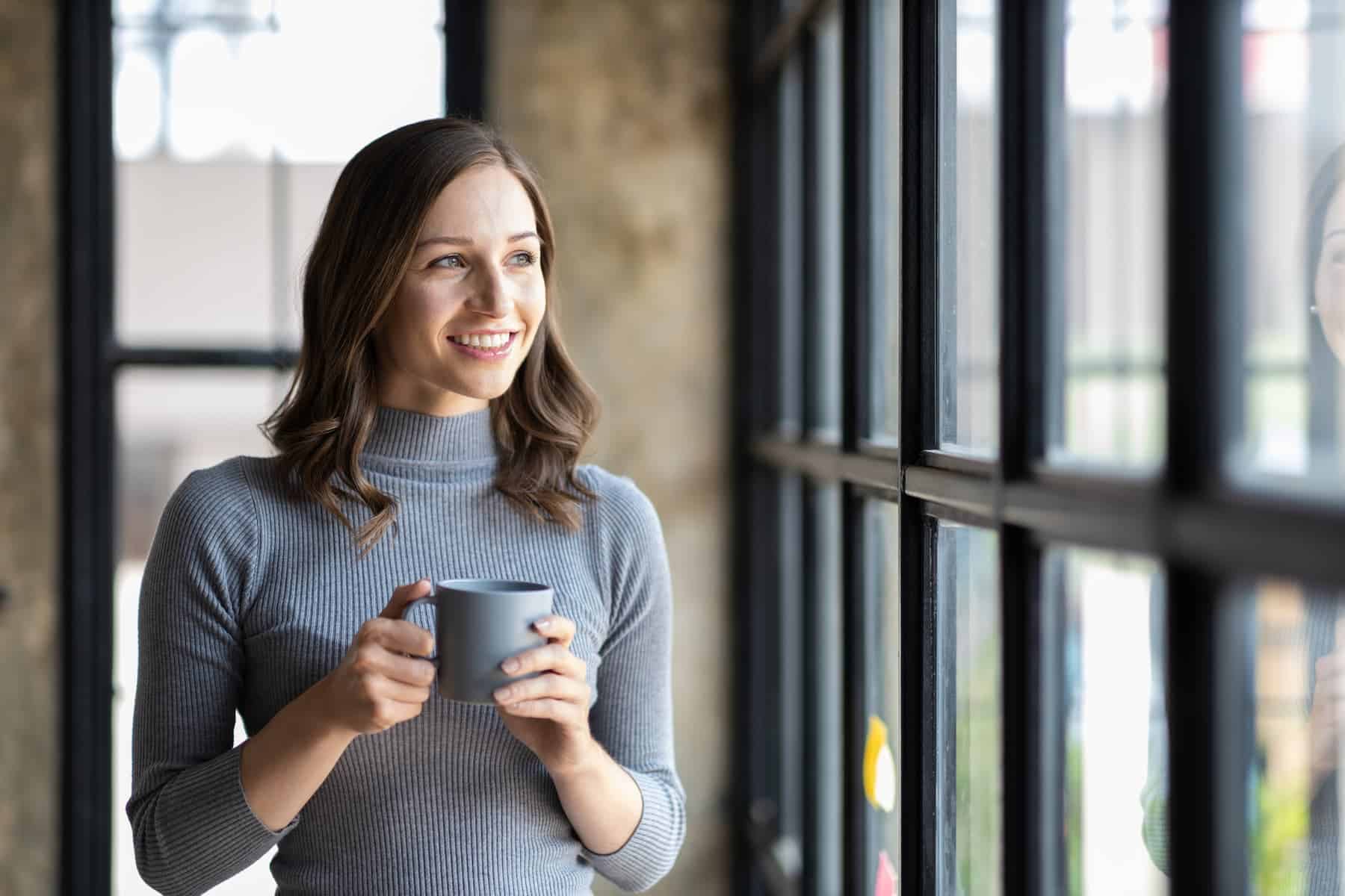 A smiling woman in a gray sweater holding a warm mug by the window, enjoying a quiet moment as part of her self-care routines.
