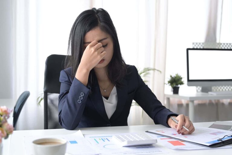 Overwhelmed businesswoman sitting at her office desk feeling exhausted and burned out from trying to achieve overly ambitious goals.