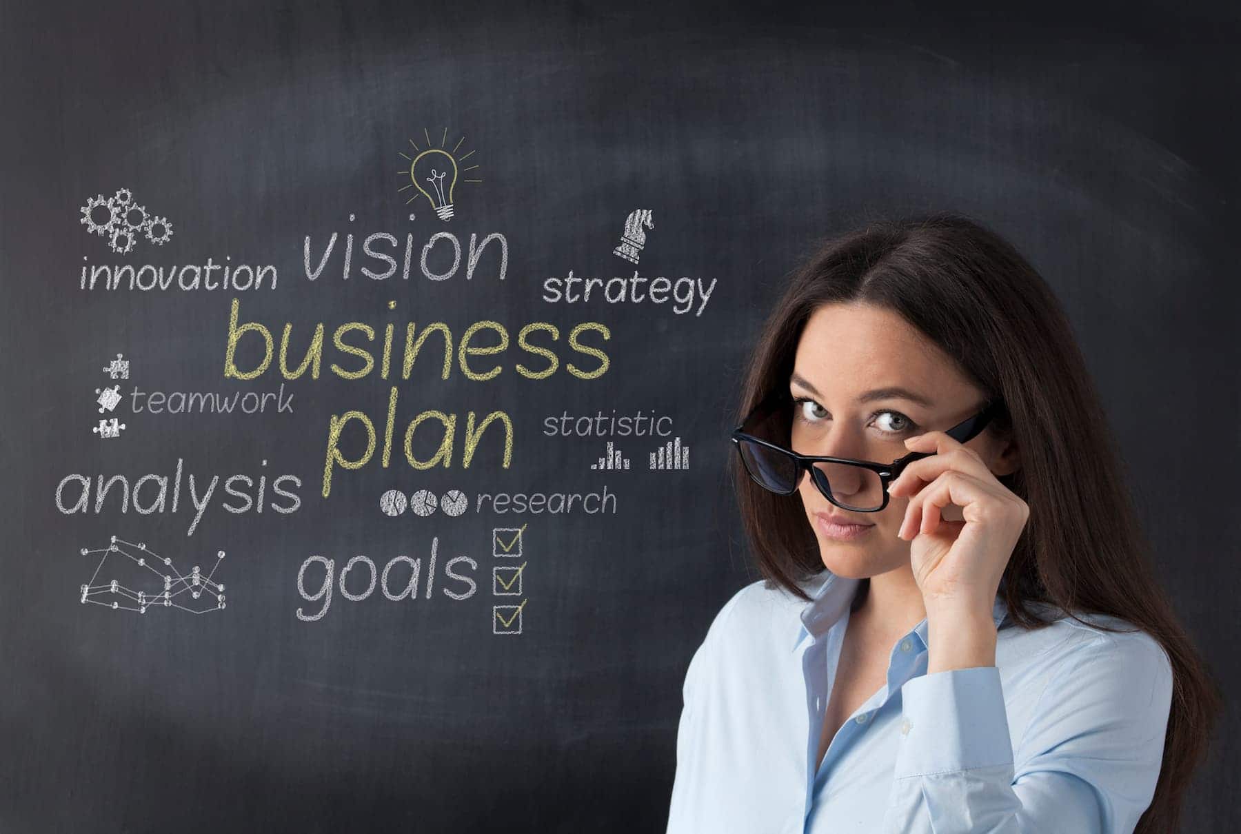 A woman in glasses stands before a chalkboard with business words like “strategy,” “vision,” and “goals.”