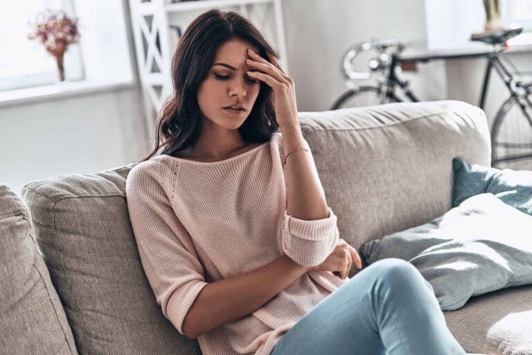 A woman sits on a couch with her hand on her forehead, looking distressed.