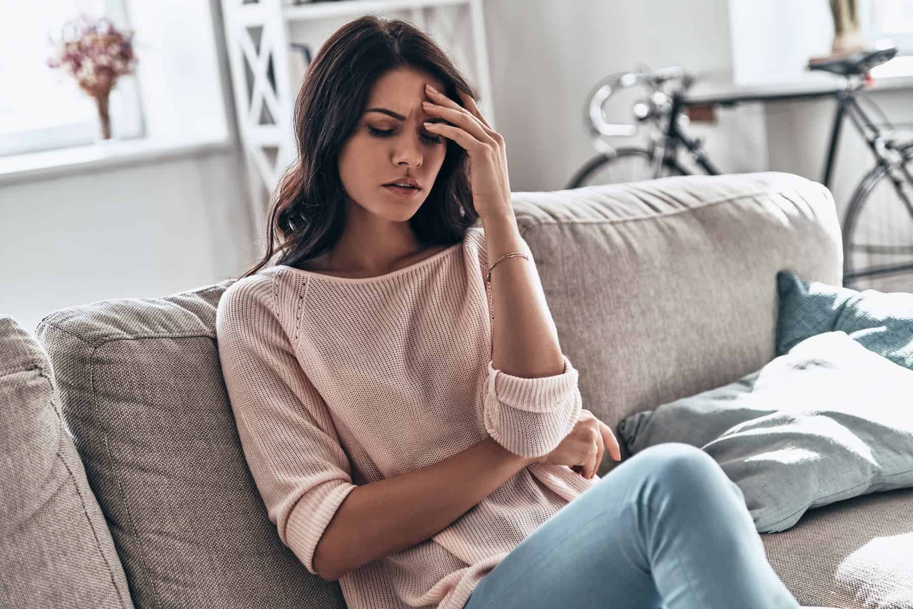 A woman sits on a couch with her hand on her forehead, looking distressed.