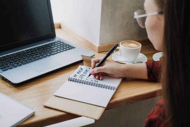 A woman in glasses writes “My Main Life Goals” in a notebook beside a laptop and coffee.