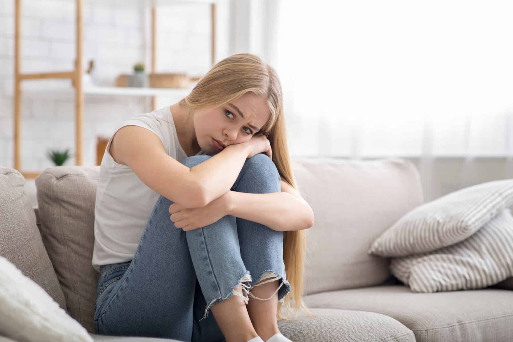 A highly sensitive woman, who has a fear of rejection, sits alone on a gray sofa, looking visibly sad.