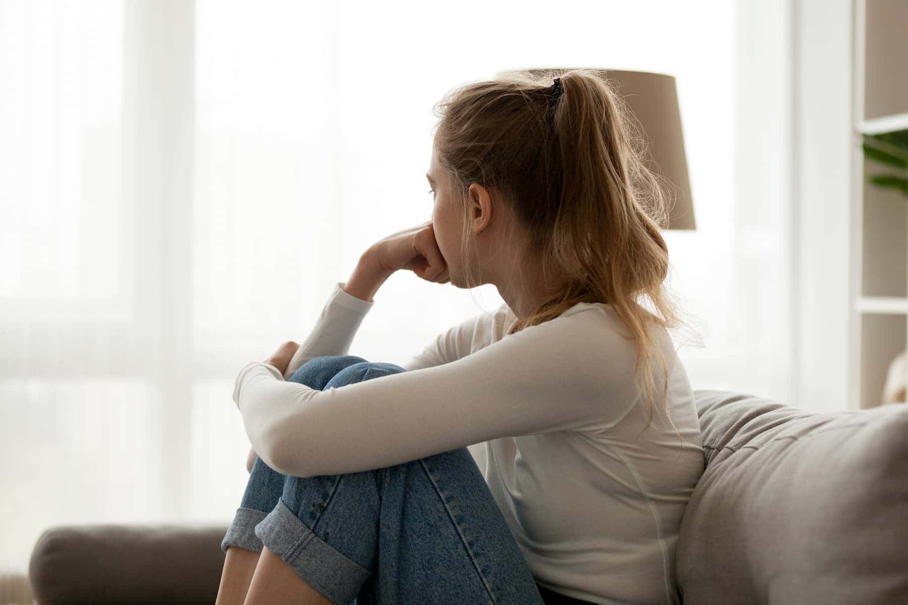 A woman sits curled up on a couch, looking out the window with her hand on her chin.