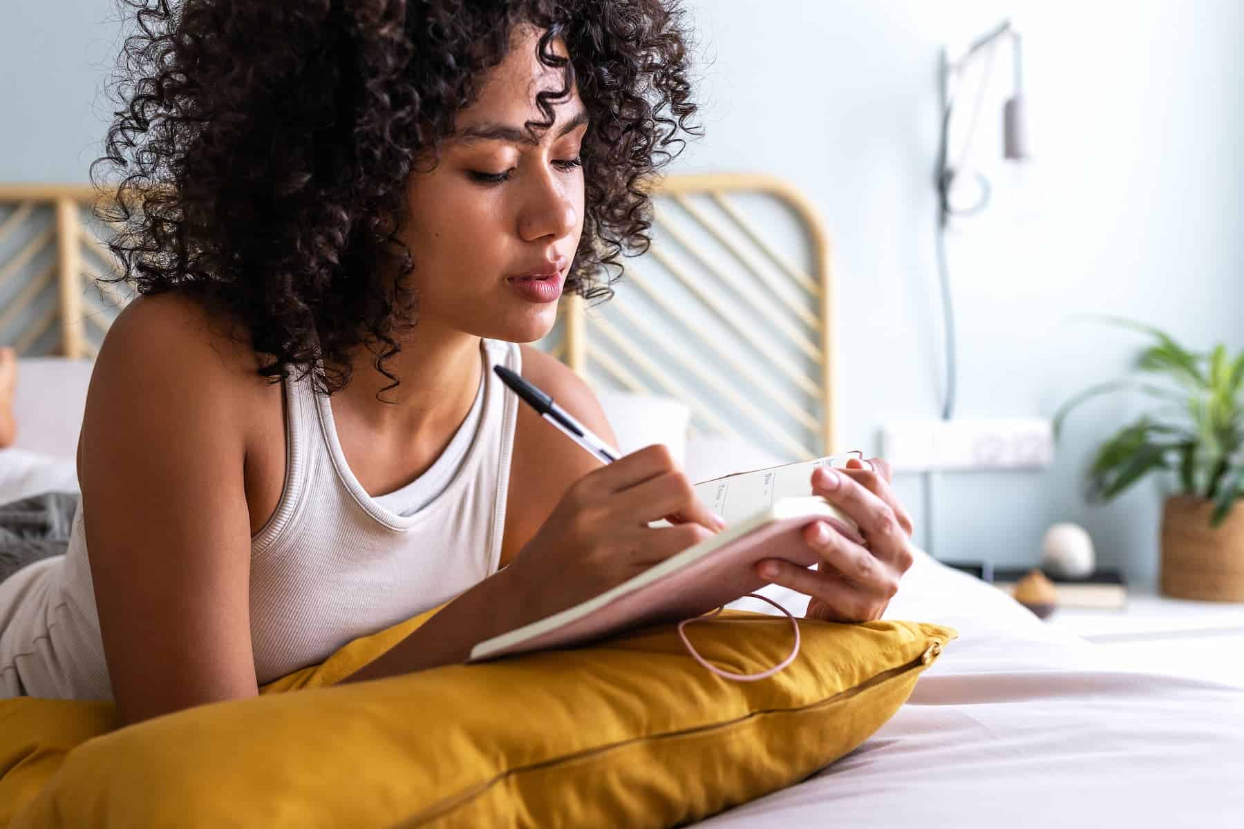 A highly sensitive introvert woman lies on her bed, with a serious expression, writing in her journal in a cozy, serene bedroom.