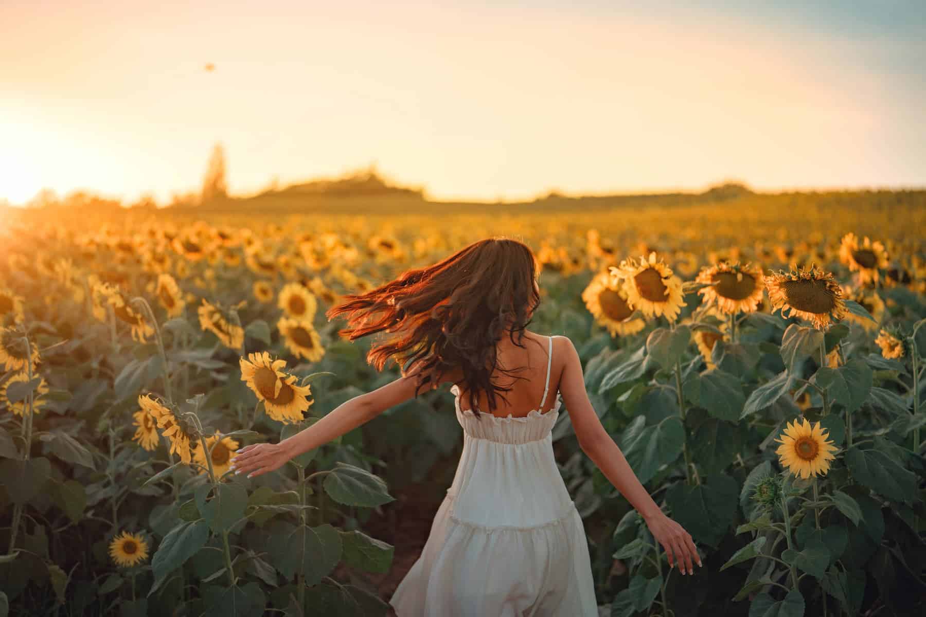 A joyful young woman in a white dress stands with her arms raised, watching the sunset over a field of sunflowers, and is starting to feel a sense of spiritual awakening.