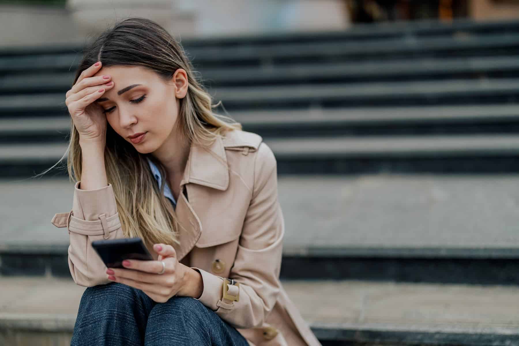 A woman sitting on the stairs, holding her phone and reading troubling news, feeling deeply distressed — a reflection of what it means to be an empath.