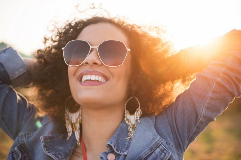 A woman wearing sunglasses is smiling in nature with the sunset behind her, noticing the subtle signs of spiritual awakening.