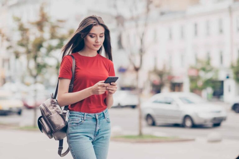 A young, beautiful girl with straight hair, wearing a casual red t-shirt and jeans, walks outside while searching for the true meaning of ambivert on her phone.