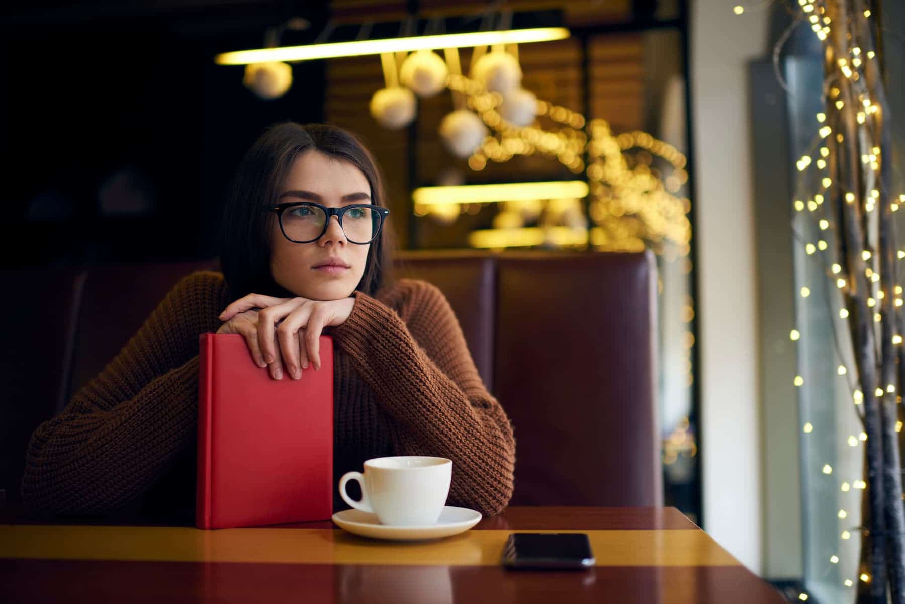 A highly sensitive woman sits in a coffee shop, her chin resting on a closed book as she enjoys her coffee, quietly reflecting on why success feels so challenging as an HSP.