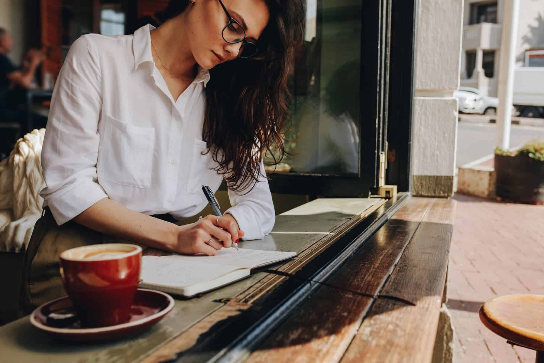 A woman sits by the café window, sipping a cappuccino as she writes personal growth plans in her notebook.