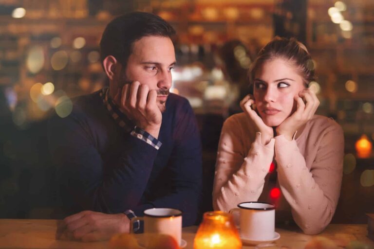 A couple in cozy sweaters sits at a dimly lit restaurant table, resting their faces on their hands and looking bored.