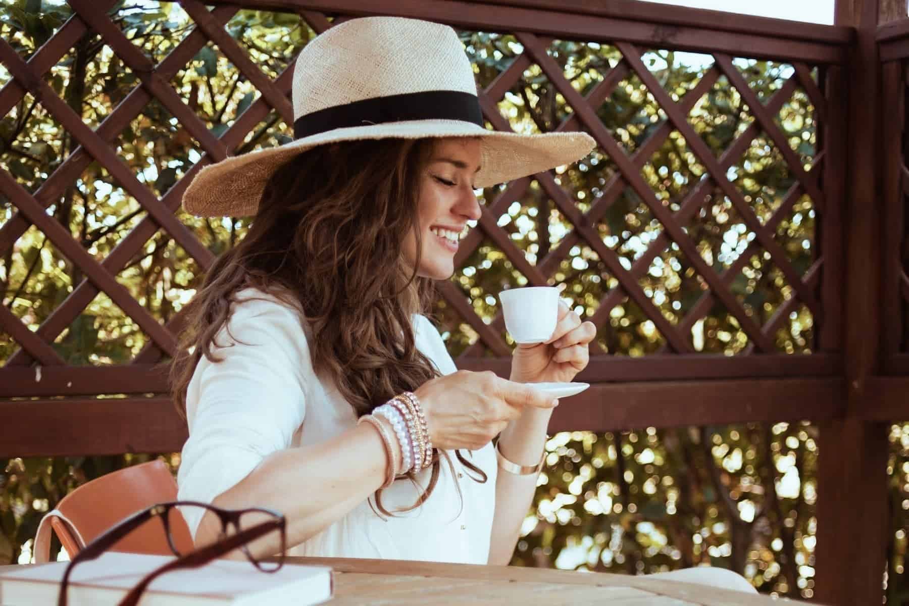 Sitting outside with a hat on, a woman savors her espresso while her book and glasses rest nearby, knowing that little things like this matter in her self-care routine.