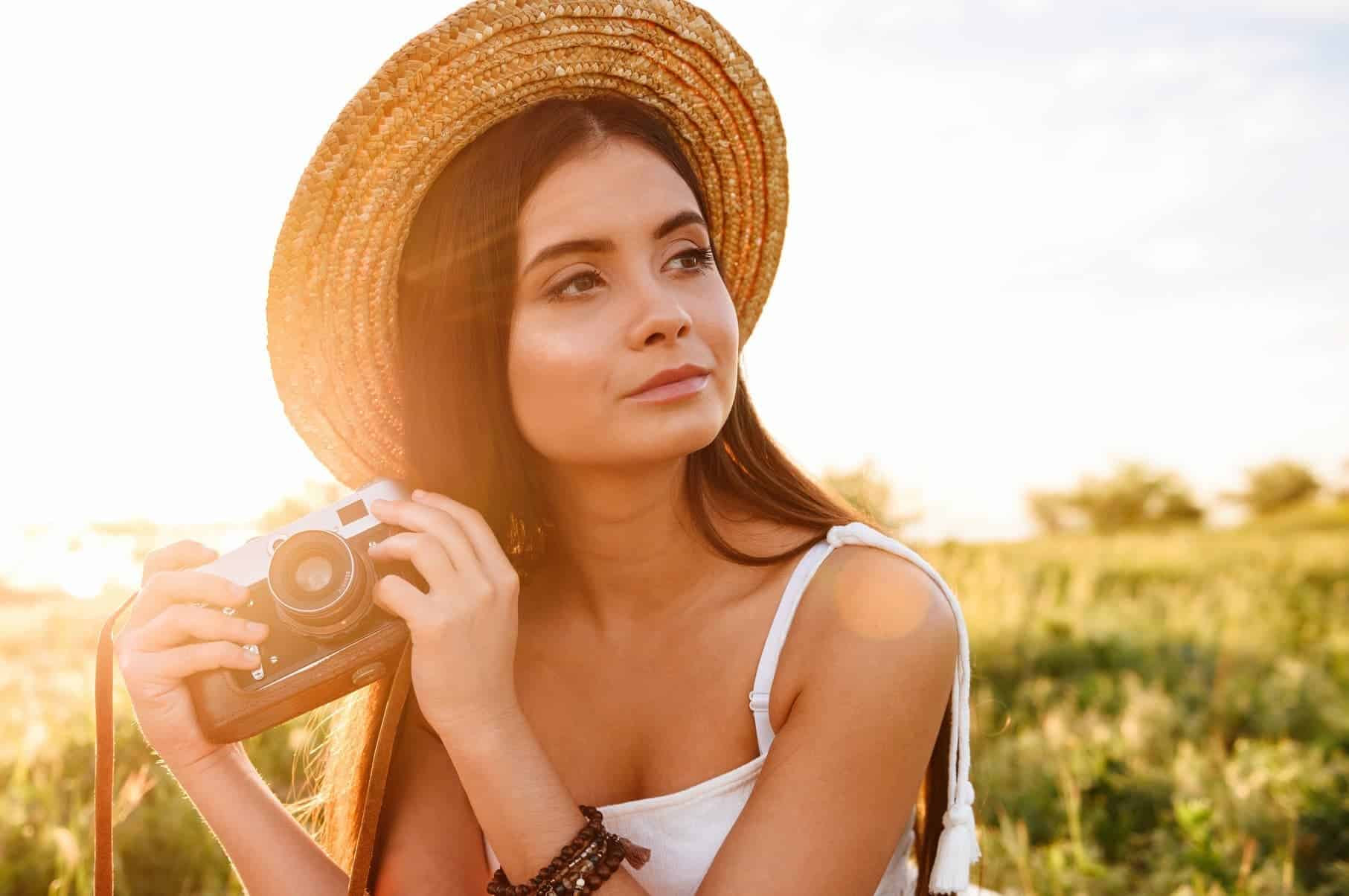 A woman wearing a hat and holding a vintage camera sits in nature with the sun shining behind her, embracing her journey of self-discovery and passion.