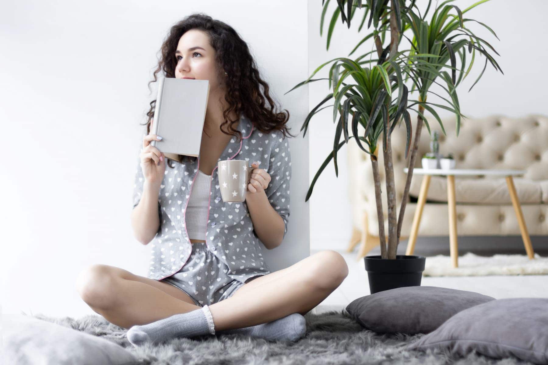 Dressed in a cute pajama set and cozy socks, a woman sits on the floor with a mug in hand, her journal and a green plant nearby, embodying the self-care essentials she values.