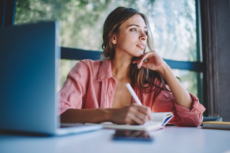 Smiling woman journaling and daydreaming, symbolizing clarity and intentional planning to create a life she loves without burnout.