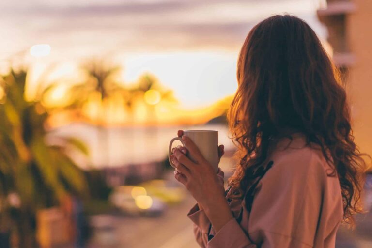 Woman sipping tea while watching a peaceful sunset from her balcony, reflecting calm and mindful daily habits for a better life.