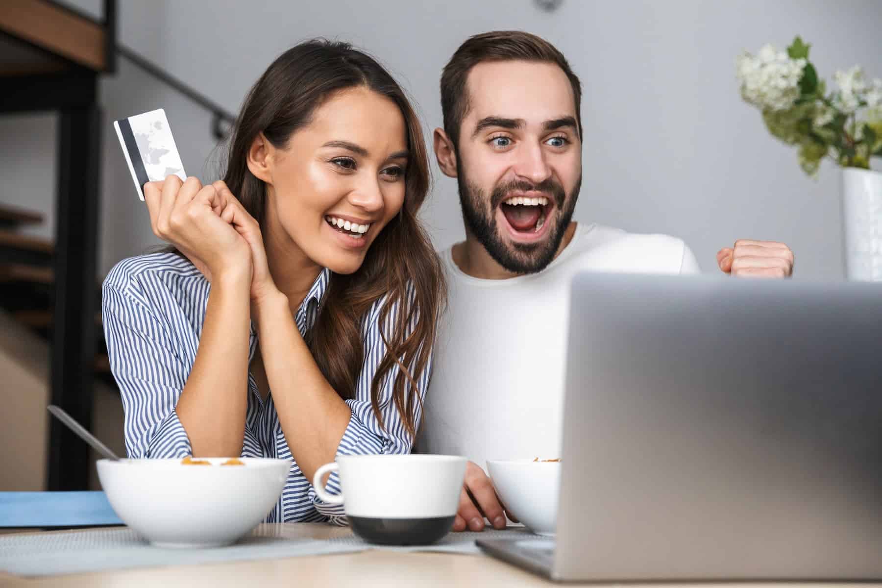 A happy couple having breakfast shop online together, with the woman holding a credit card and the man looking excited about the amazing Amazon Prime deals.